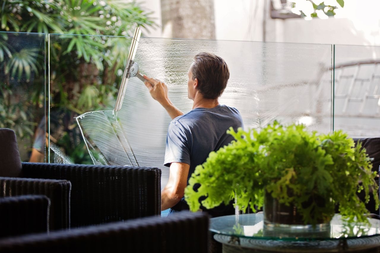 Free A man cleaning a glass barrier outdoors with a squeegee, surrounded by greenery. Stock Photo