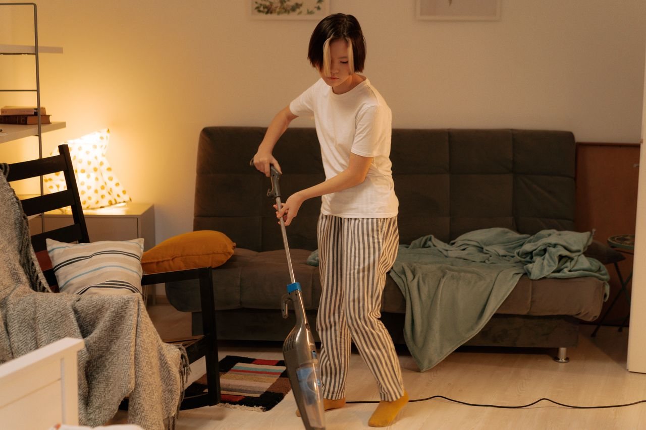 Free Asian woman cleaning a messy living room at home with a vacuum cleaner, creating a tidy space. Stock Photo
